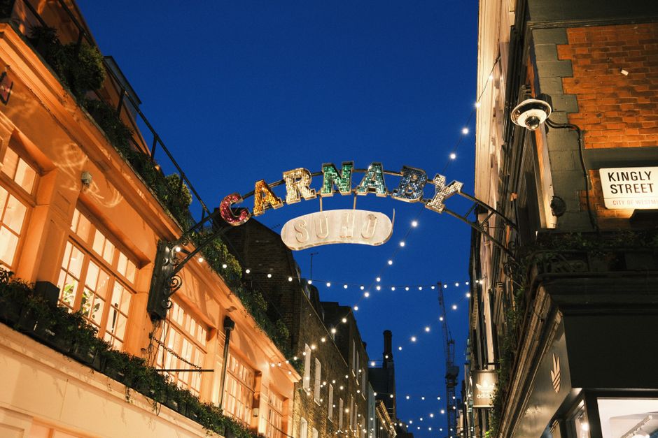 Nighttime exterior scene showing the illuminated sign of a Soho street named Carnaby with a smaller sign beneath it reading 'Studio'. The street is lined with buildings featuring large windows, some w